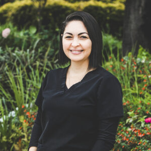 A woman with a fair complexion, shoulder-length, dark straight black hair and wearing a black medical outfit is seen standing in front of an urban garden on a bright summer day