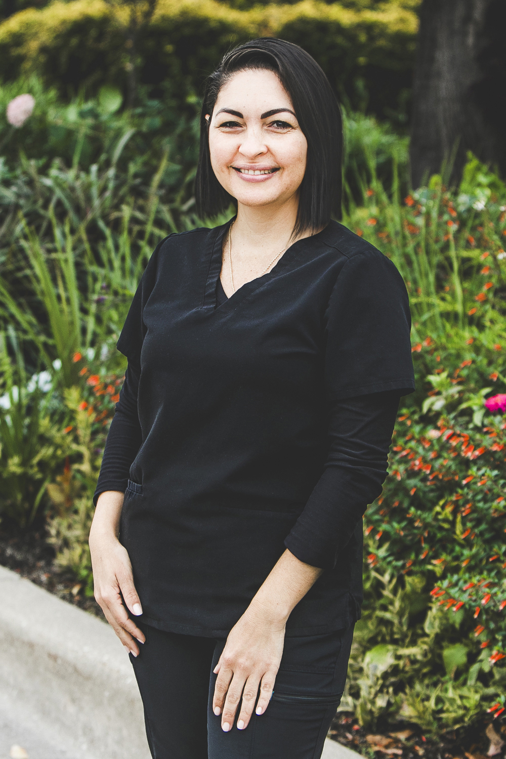 A woman with a fair complexion, shoulder-length, dark straight black hair and wearing a black medical outfit is seen standing in front of an urban garden on a bright summer day