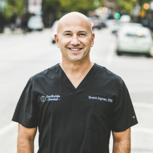 A man with a medium complexion and shaved head, and wearing a modern, dark-grey medical outfit stands in front of a bustling city street on a bright, sunny day