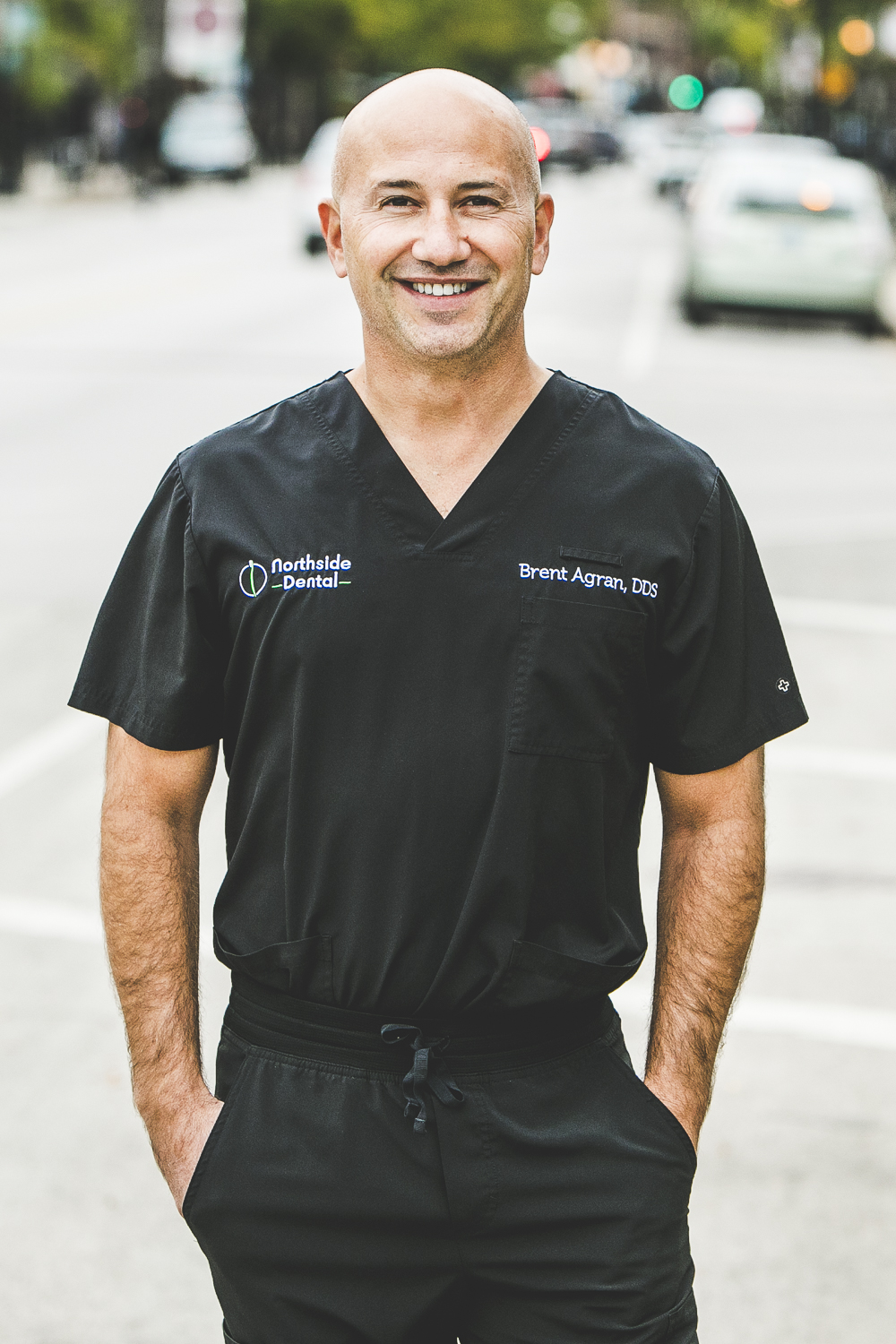 A man with a medium complexion and shaved head, and wearing a modern, dark-grey medical outfit stands in front of a bustling city street on a bright, sunny day