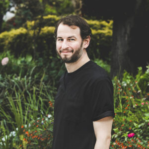 A man with a fair, complexion, short, wavy brown hair and brown beard with flecks of grey and wearing a black medical outfit is seen standing in front of an urban garden on a bright summer day