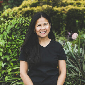 A woman with a medium complexion, long, dark straight black hair and wearing a black medical outfit is seen standing in front of an urban garden on a bright summer day