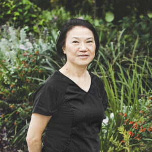 A woman with short, straight, neck-length black hair and wearing a black medical outfit is seen standing in front of an urban garden on a bright summer day