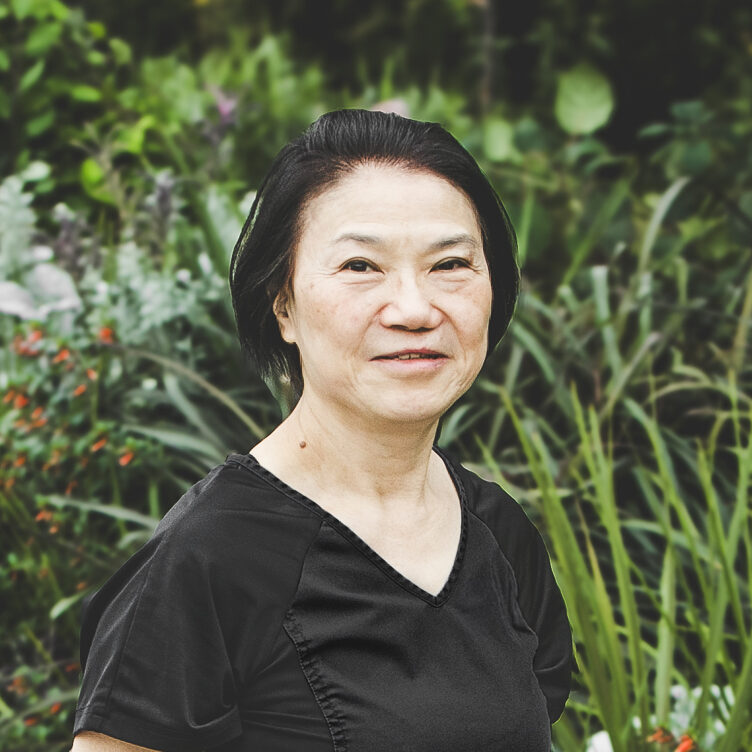 A woman with short, straight, neck-length black hair and wearing a black medical outfit is seen standing in front of an urban garden on a bright summer day