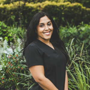 A woman with a medium complexion and long, black wavy hair and wearing a dark grey medical outfit is seen standing in front of an urban garden on a bright summer day