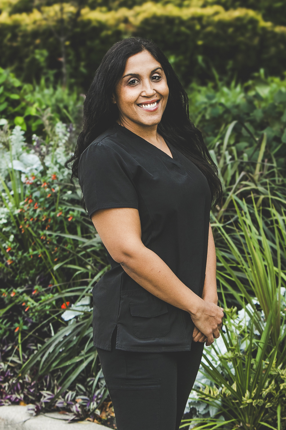 A woman with a medium complexion and long, black wavy hair and wearing a dark grey medical outfit is seen standing in front of an urban garden on a bright summer day