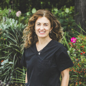 A woman with a fair complexion with slightly-curly shoulder-length brown hair with highlights wearing a dark-grey medical outfit is seen standing in front of an urban garden on a bright summer day