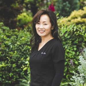 A woman with long, wavy, black and brown hair and wearing a dark-grey medical outfit with the label Dr. Jin embroidered to the pocket is standing in front of a lush city garden on a bright summer day