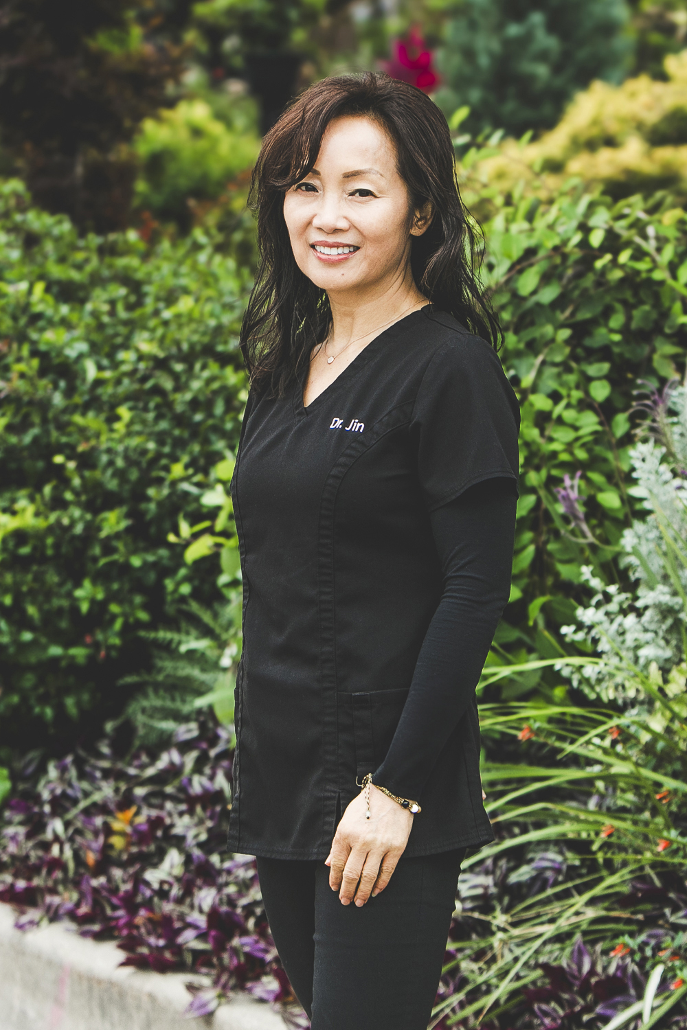 A woman with long, wavy, black and brown hair and wearing a dark-grey medical outfit with the label Dr. Jin embroidered to the pocket is standing in front of a lush city garden on a bright summer day