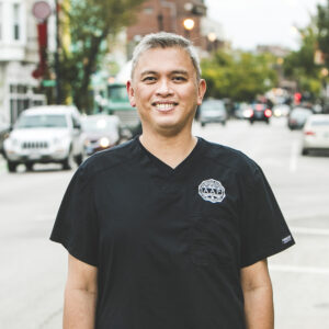 A man with a medium complexion and salt and pepper hair is smiling while standing on a busy city street on a bright summer day