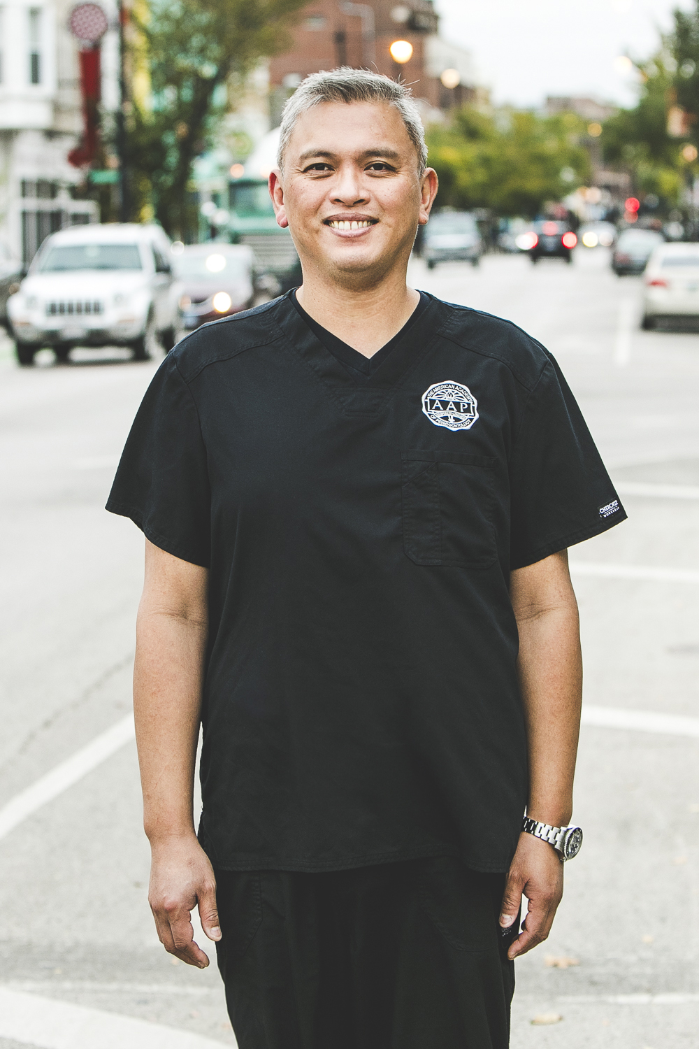 A man with a medium complexion and salt and pepper hair is smiling while standing on a busy city street on a bright summer day