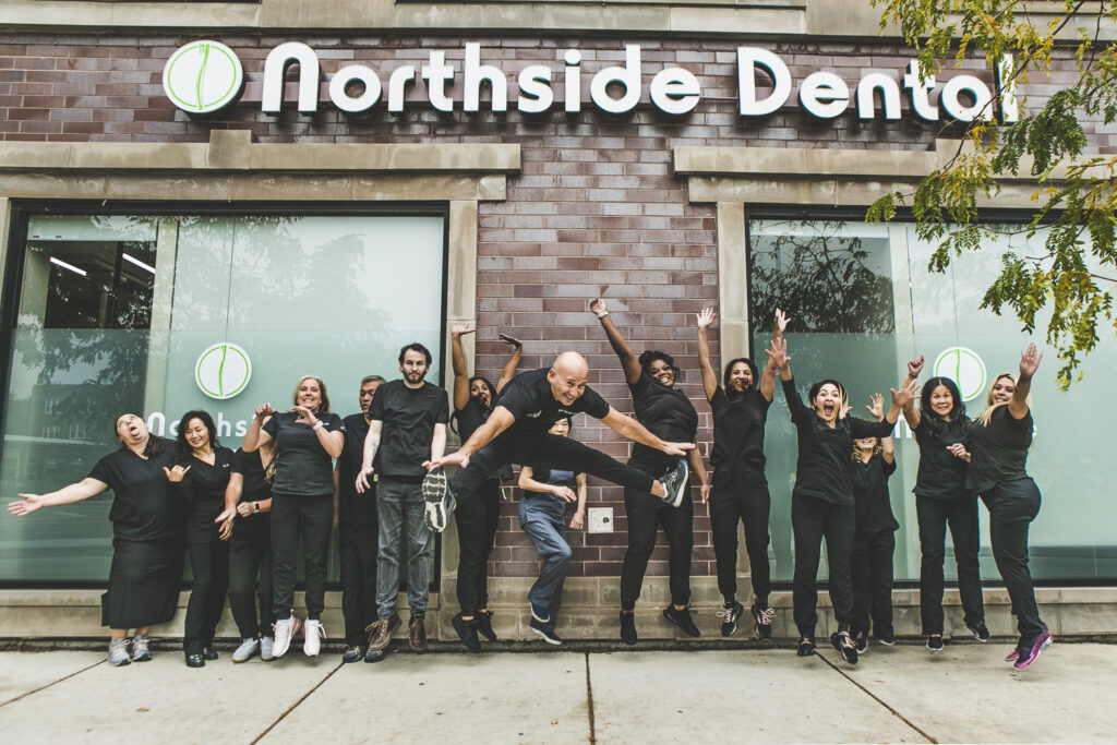 A large group photo. A row of dentists and dental technicians & assistants, all in black or dark-grey, modern medical outfits are having fun by jumping up in front of the camera at once for an action shot. They're outside a modern-looking, brick office façade, just underneath a large Northside Dental exterior sign