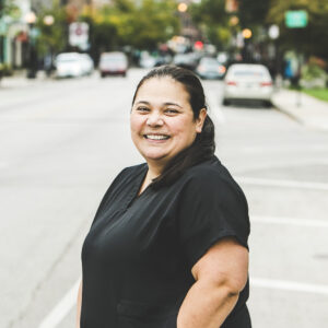 A woman with a light-tan complexion with long, straight black hair pulled into a pony tail and wearing a black medical outfit is seen standing outside in front of a bustling city street on a bright, summer day