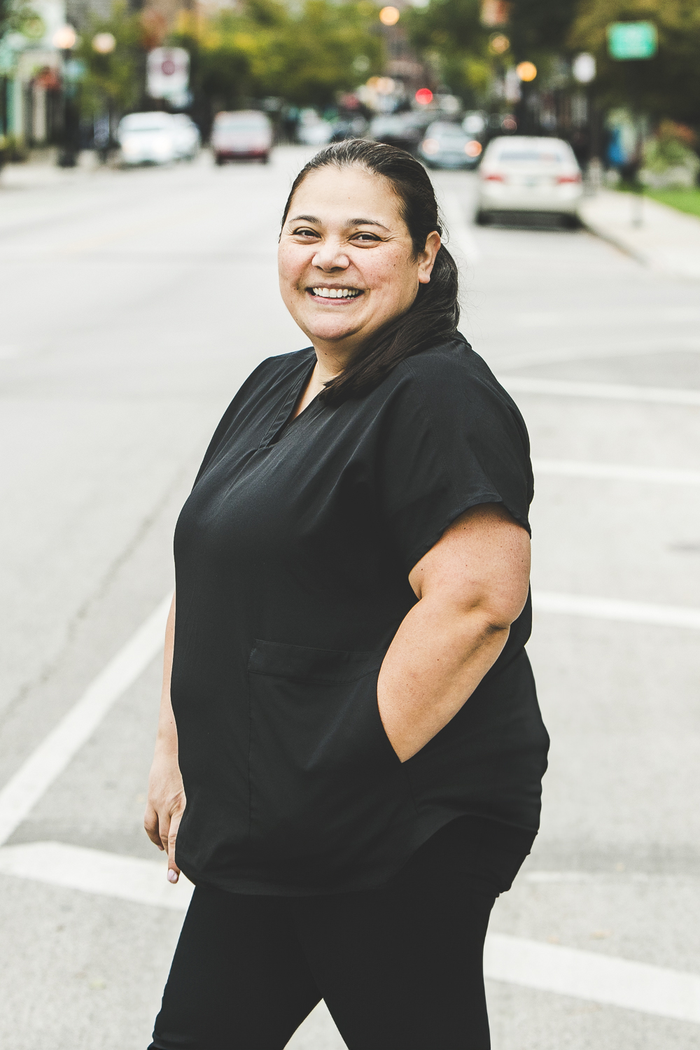 A woman with a light-tan complexion with long, straight black hair pulled into a pony tail and wearing a black medical outfit is seen standing outside in front of a bustling city street on a bright, summer day