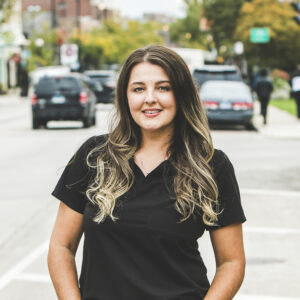 A woman with a light-tan complexion with long, wavy brown hair with highlights wearing a black medical outfit is seen standing outside in front of a bustling city street on a bright, summer day