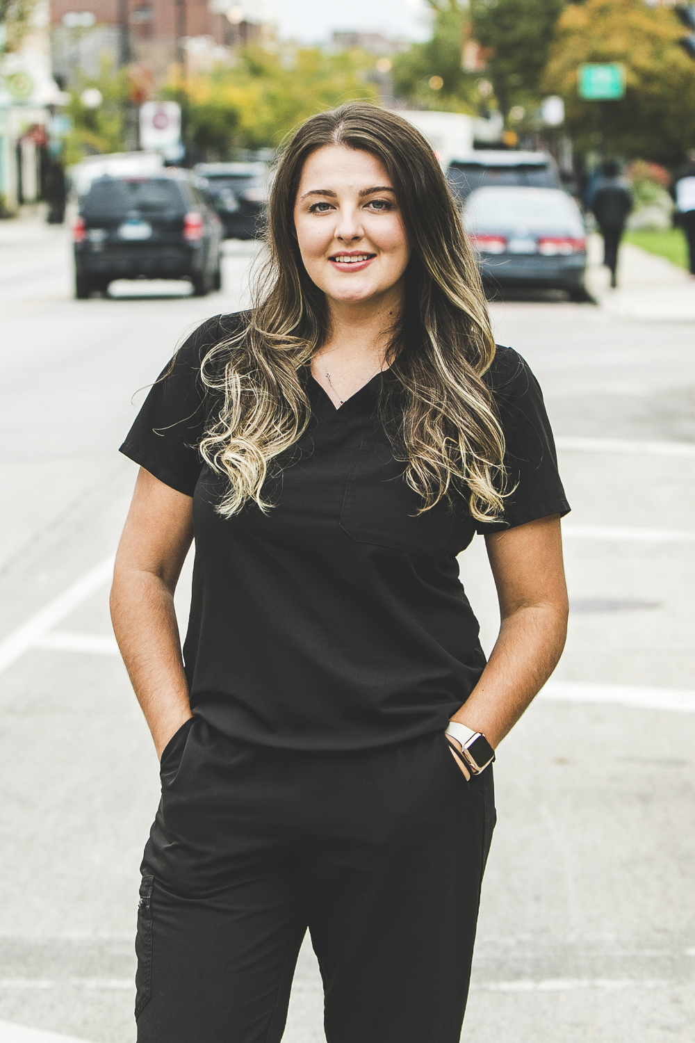 A woman with a light-tan complexion with long, wavy brown hair with highlights wearing a black medical outfit is seen standing outside in front of a bustling city street on a bright, summer day