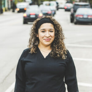 A woman with a tan complexion and medium-length, curly, sandy blonde hair, wearing a black headband and black medical outfit is seen standing outside in front of a bustling city street on a bright, summer day