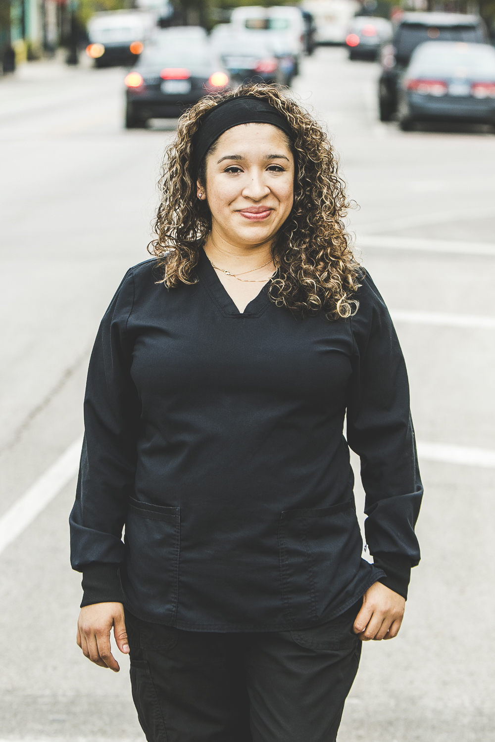 A woman with a tan complexion and medium-length, curly, sandy blonde hair, wearing a black headband and black medical outfit is seen standing outside in front of a bustling city street on a bright, summer day