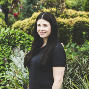 A woman with a a fair complexion and long, dark straight black hair and wearing a black medical outfit is seen standing in front of an urban garden on a bright summer day