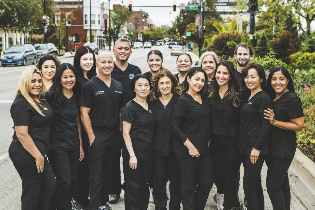 A group of dentists and dental technicians in dark-colored medical outfits are standing on a city street mid-day and smiling.
