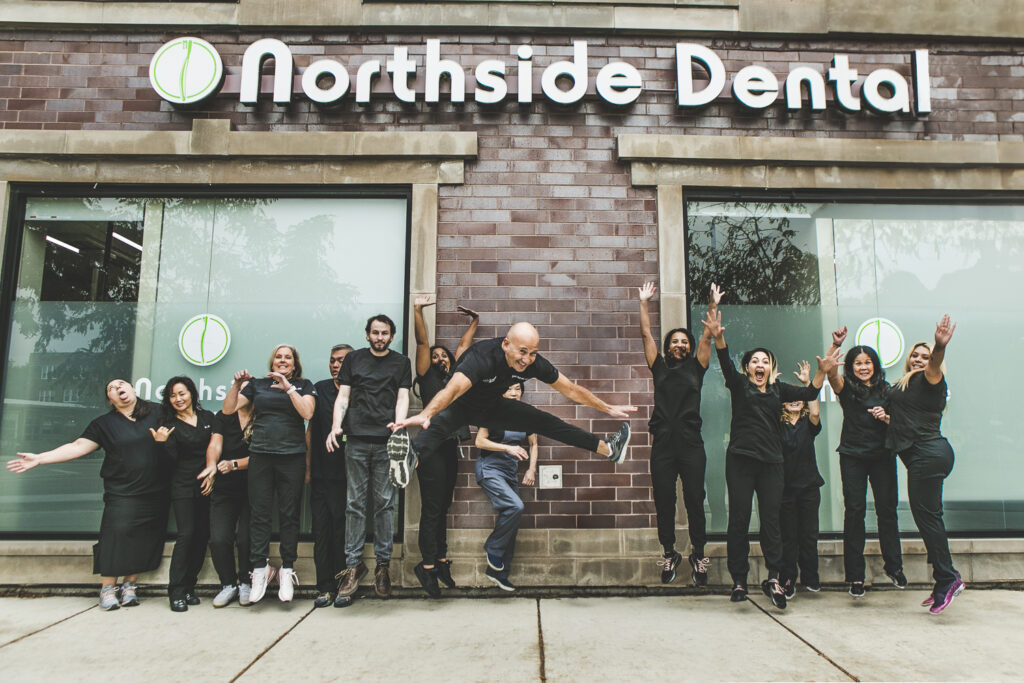 A large group photo. A row of dentists and dental technicians & assistants, all in black or dark-grey, modern medical outfits are having fun by jumping up in front of the camera at once for an action shot. They're outside a modern-looking, brick office façade, just underneath a large Northside Dental exterior sign