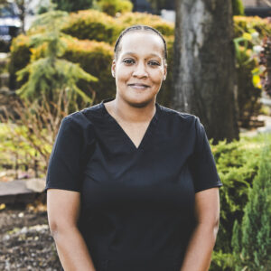 A woman with a medium dar complexion and wearing her hair in a short, tightly-braided style is seen in a park setting wearing a black-colored medical outfit on a bright day.