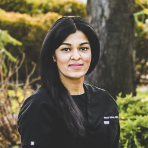 A woman with a tan complexion and long, dark, straight hair is seen wearing a full black medical outfit and standing in a park setting on a bright day.
