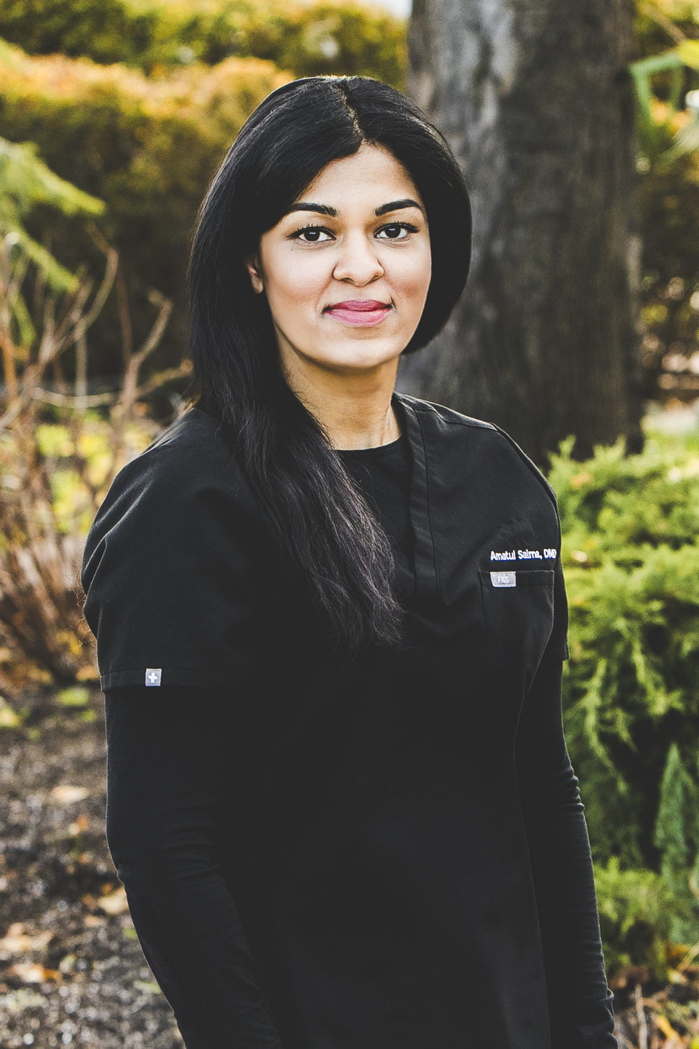 A woman with a tan complexion and long, dark, straight hair is seen wearing a full black medical outfit and standing in a park setting on a bright day.